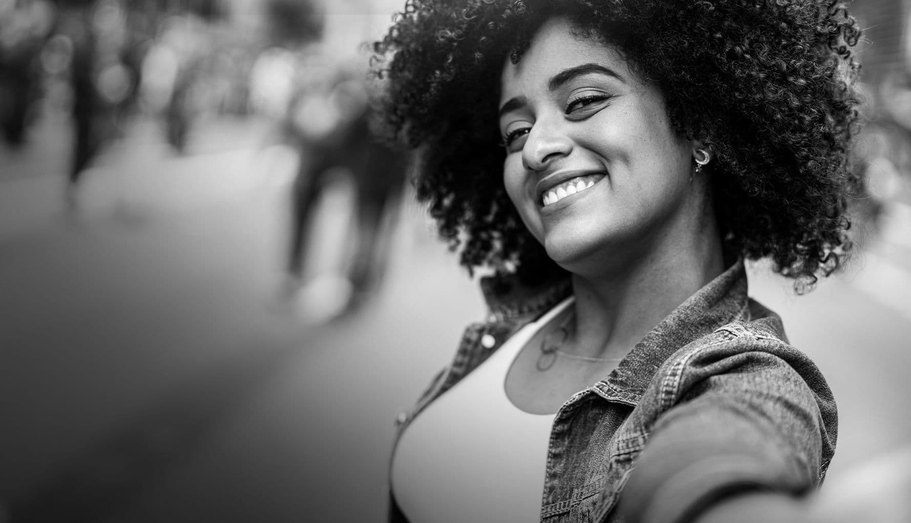Donna sorridente con capelli ricci e giacca di jeans che si fa un selfie, con una strada sfocata sullo sfondo